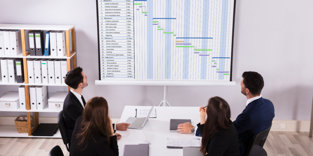 A group of people in an office looking at a Gantt chart being projected onto a screen.  4 people look at the large chart looking in deep thought.